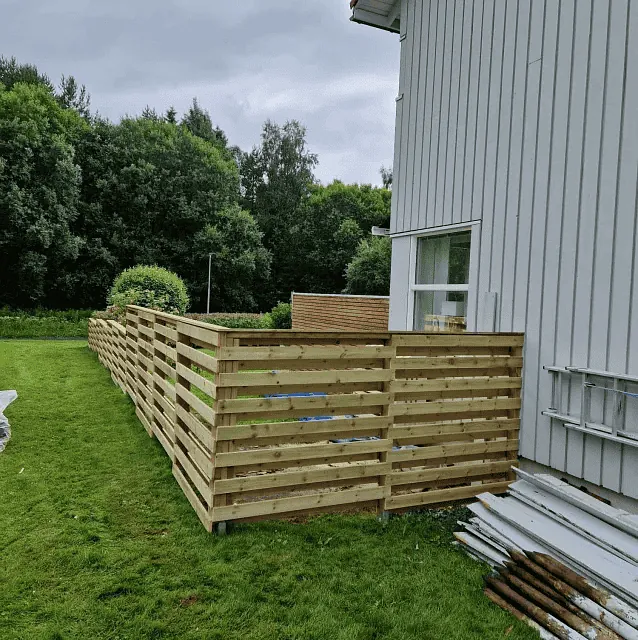 Nyoppført tre-gjerde langs en hvit bolig med grønt gress og skog i bakgrunnen.